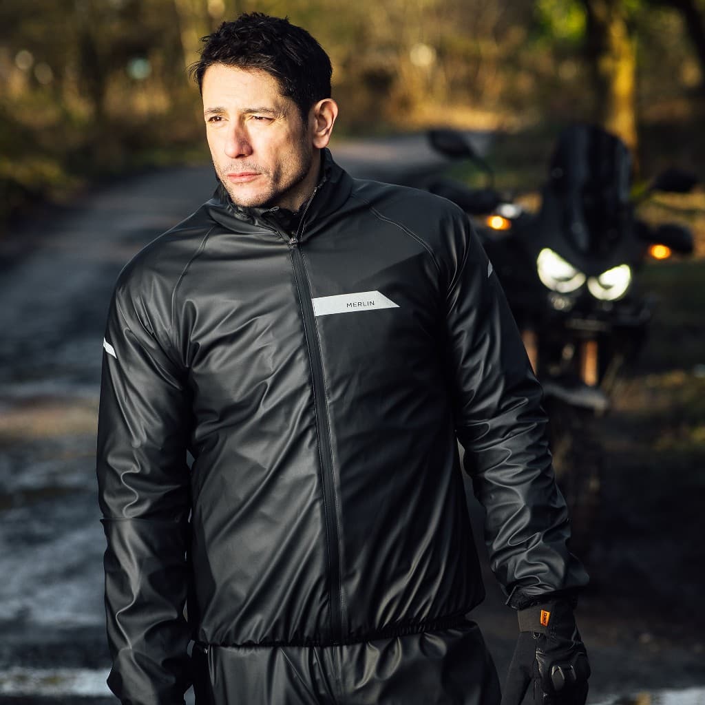 Rider wearing a Merlin waterproof motorcycle rain jacket with reflective panels standing in front of a motorcycle on a winter road