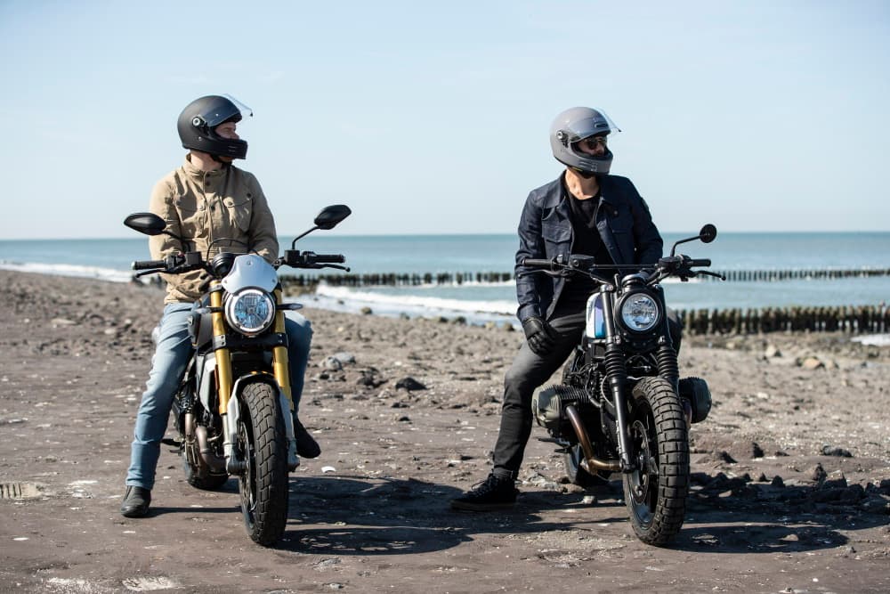 Two motorcyclists wearing full-face helmets and retro riding gear on a beach with custom scrambler and cafe racer motorcycles